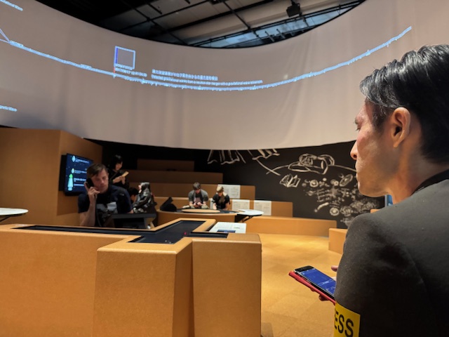 ベンチにすわりながら、映像展示をたのしんでいるしらとりさんの後ろ姿を写した写真。右手にわアシストアプリプラスをひらいたスマホをもっています