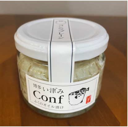 ふぐのオイル漬け「Conf」の瓶詰の写真。瓶は透明なガラス製で、なかにはオイル漬けされたふぐの身が見えます。瓶の蓋は白色で、中央に細い白色のラベルが貼られており、「コンフプレーン」と縦書きで印字されています。その下の正面には白いラベルが貼られており、上部に「ハカタいづみ」と日本語が書かれ、そのしたに大きな文字で「Conf」と印字されています。さらに下には「ふくのオイル漬け」と書かれ、右下にはふぐのイラストと赤い印鑑がデザインされています