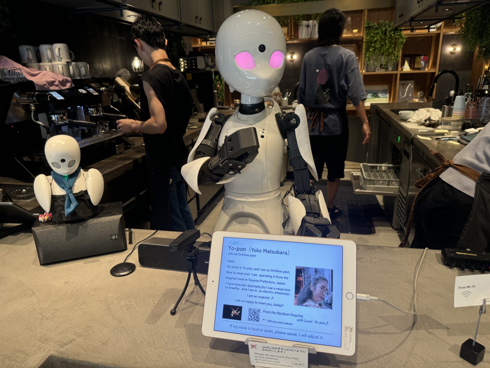 分身ロボットカフェドーンバージョンベータの受付でお客様を迎えている120センチの大きなオリヒメの写真。右手を胸に当てて「かしこまりました」のポーズをしている。目の色は、パイロットが自分で選んだピンクいろ