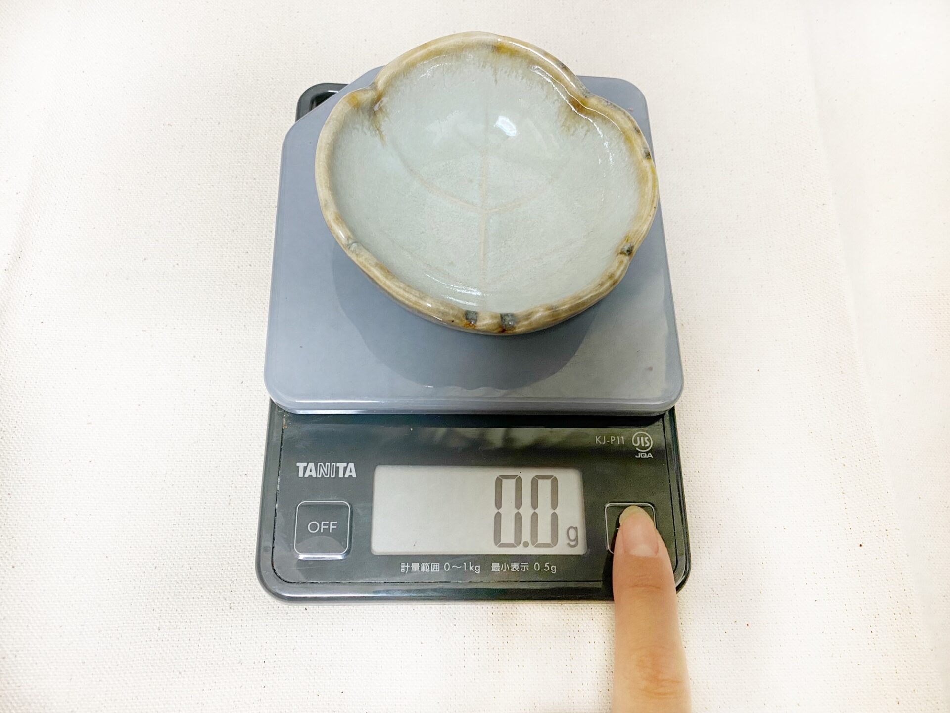 クッキングスケールの計量皿にお皿をのせたまま、指でボタンをおしている写真。お皿の重さが差し引かれて０．０グラムと表示されています
