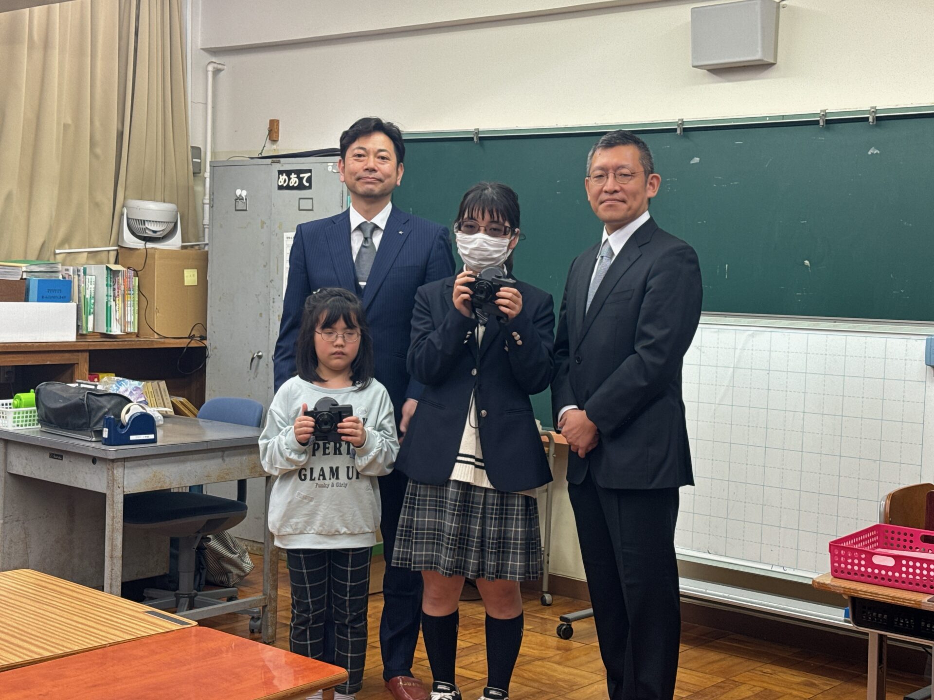 もうがっこうの教室内で、贈呈した網膜投影カメラキットを手にした女子生徒2名と、ソニーのおおしままさあきさんとQDレーザのみやうちひろのりさんの写真