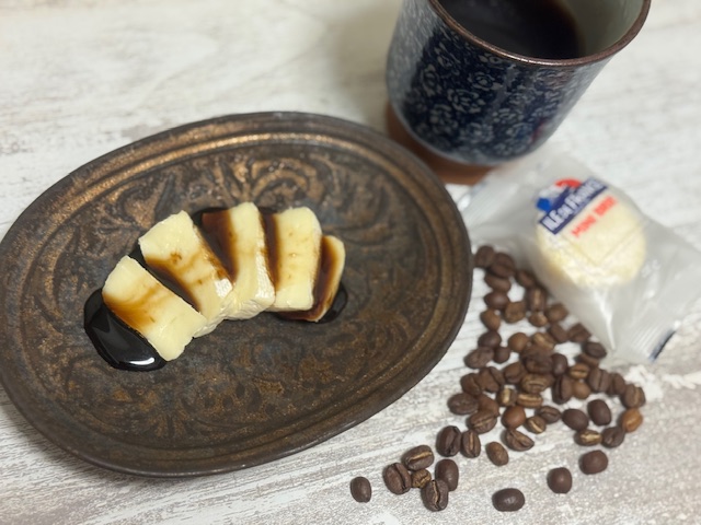 だえんがたのちゃいろっぽいおさらに、たてに薄くカットしたミニブリーをならべ、そのうえにくろみつをかけたものがのっている写真。まわりには青いマグカップにハイッタブラックコーヒーとコーヒー豆、ミニブリーの個包装のパッケージがひとつ映り込んでいます