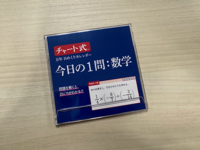 プラスチックの薄いせいほうけいのケースにハイッタひめくりカレンダーの表紙の写真。こいブルーの背景に白い文字、参考書とおなじフォントで「チャート式ばんねんひめくりカレンダー、今日のいちもん：数学」とかかれています。もんだいをとくとひにちがわかる！？とかかれており、となりにサンプル問題が。よんぶんのいち、かける、かっこマイナスななぶんのろくかっことじ、わる、マイナスじゅうよんぶんのさんかっことじと数式でかかれています たとえが古くて年代がばれそうですが、CDのマキシシングルのような、とうめいのプラスチックのケースに一枚ずつハイっています