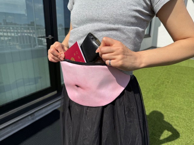 屋外でポーズをとる女性の写真。人物の顔の部分がぼかしで隠されていますが、これ、わたしです。グレーのTシャツに黒いスカートを着用し、おなかが少し見えているようにみえるのがこのウエストポーチです。そしてハンソデなので、にのうでを目立たせないようなポージングに必死です。背景には建物や手すり、空が写っており、人工芝のようなものが敷かれた場所に立っています。親会社の屋上で撮影した写真です
