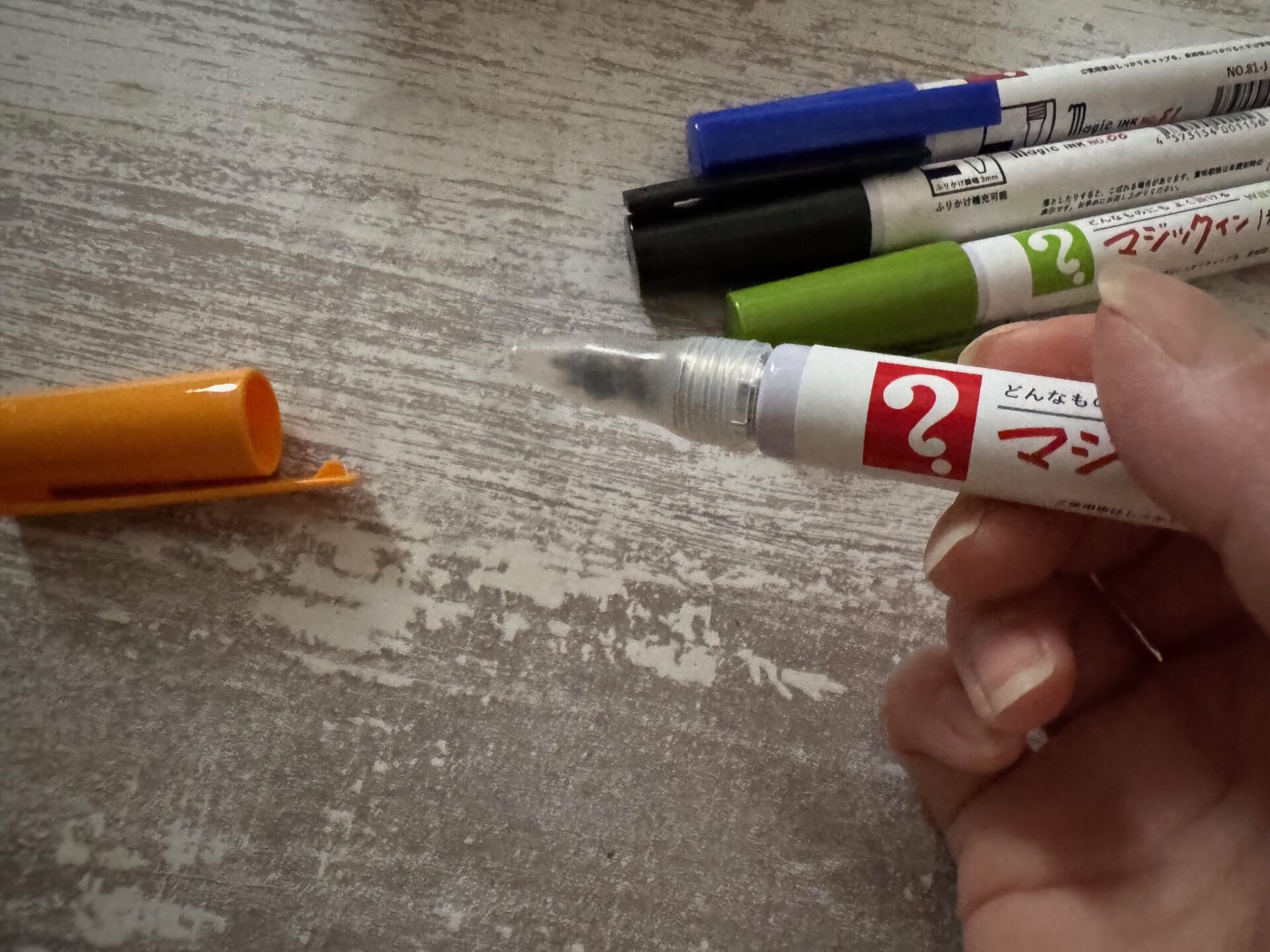 キャップをとった、くしかつあじのマジックインふりかけを、右手でもっているようすを写した写真。ペンのさきのほうをふると、ふりかけがでてくるしくみです。背後にわ、あお、くろ、みどりのペンもうつりこんでいます