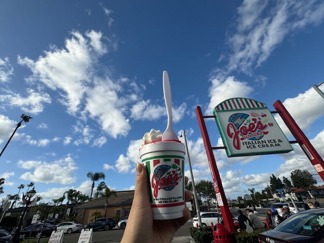 ジョーズイタリアンアイスのお店のカンバンのまえでカップにハイッタアイスを手に持った写真。背景にはカリフォルニアの青空がひろがっています