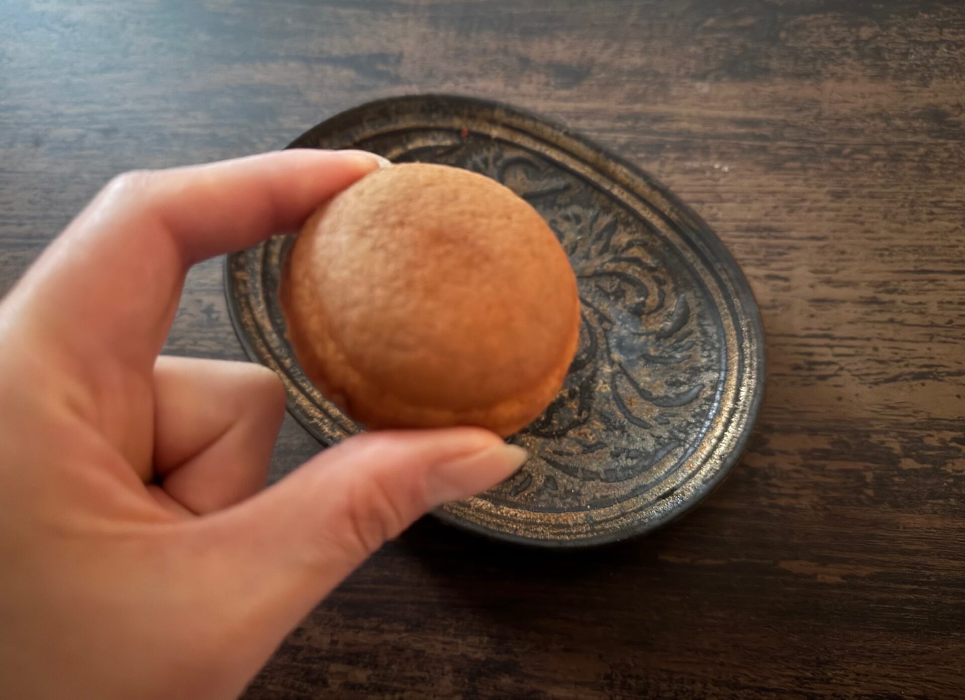 ひだりてのおやゆびとひとさしゆびで、まるくてちいさなどらやきのようなかたちのにんぎょうやきをもった写真。背後にわ茶色とゴールドのおさらも映り込んでいます