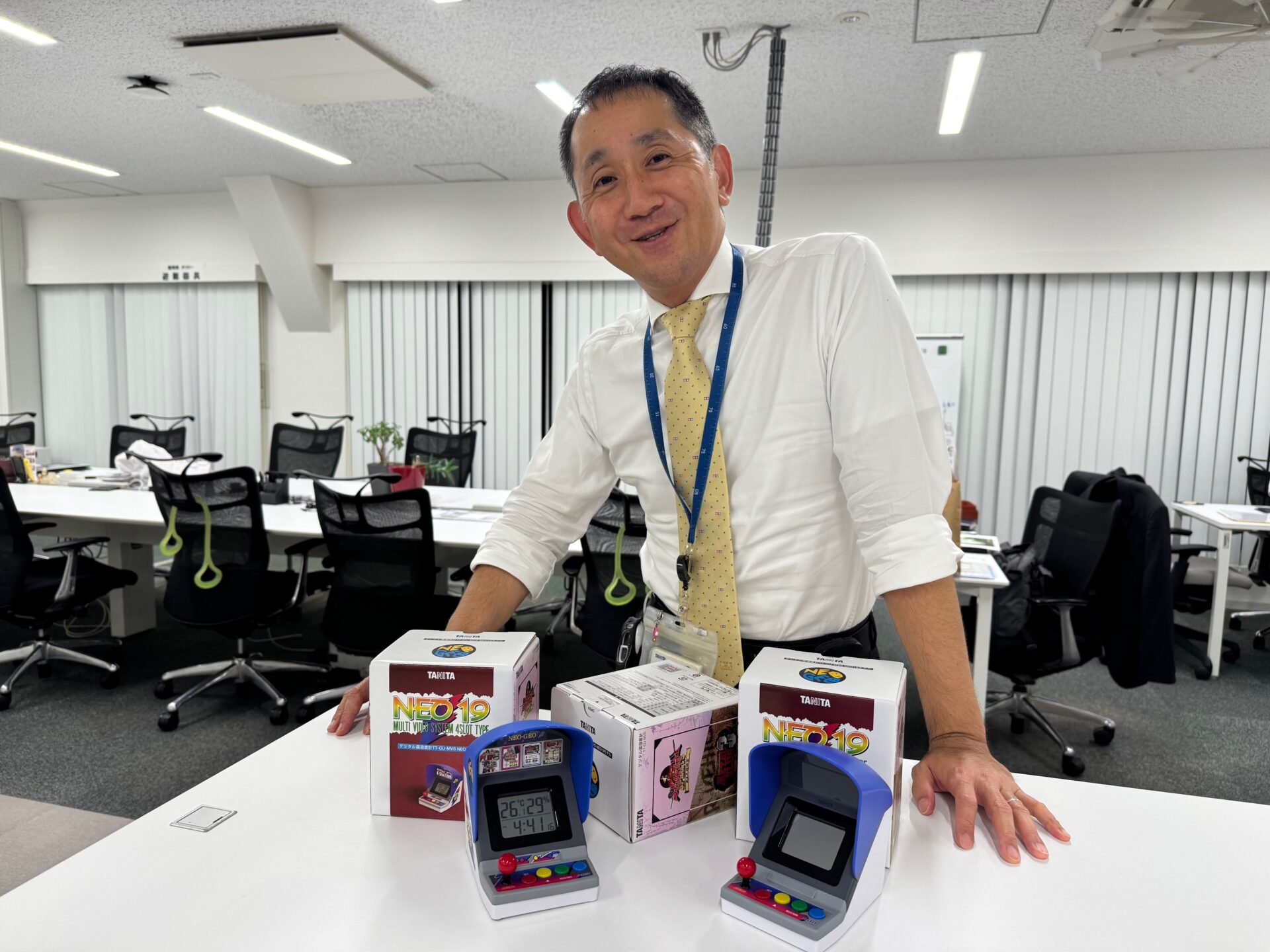 タニタのたにだせんり社長の写真。笑顔、というかドヤがおで両手をですくに置いてこちらを見つめています。デスクのうえにわ、ゲーム会社とコラボのゲームきょうたいのかたちをしたおんしつどけいがふたつ、パッケージのハコがみっつ、うすいきいろのネクタイわ、よくみるとタミヤのロゴがところどころに描かれています。でもいちばんのポイントわ、しゃいんしょうがついたネックストラップにつけられたちいさなカードキャプターさくらのチャームです。ジャンルはちがえどオタクです
