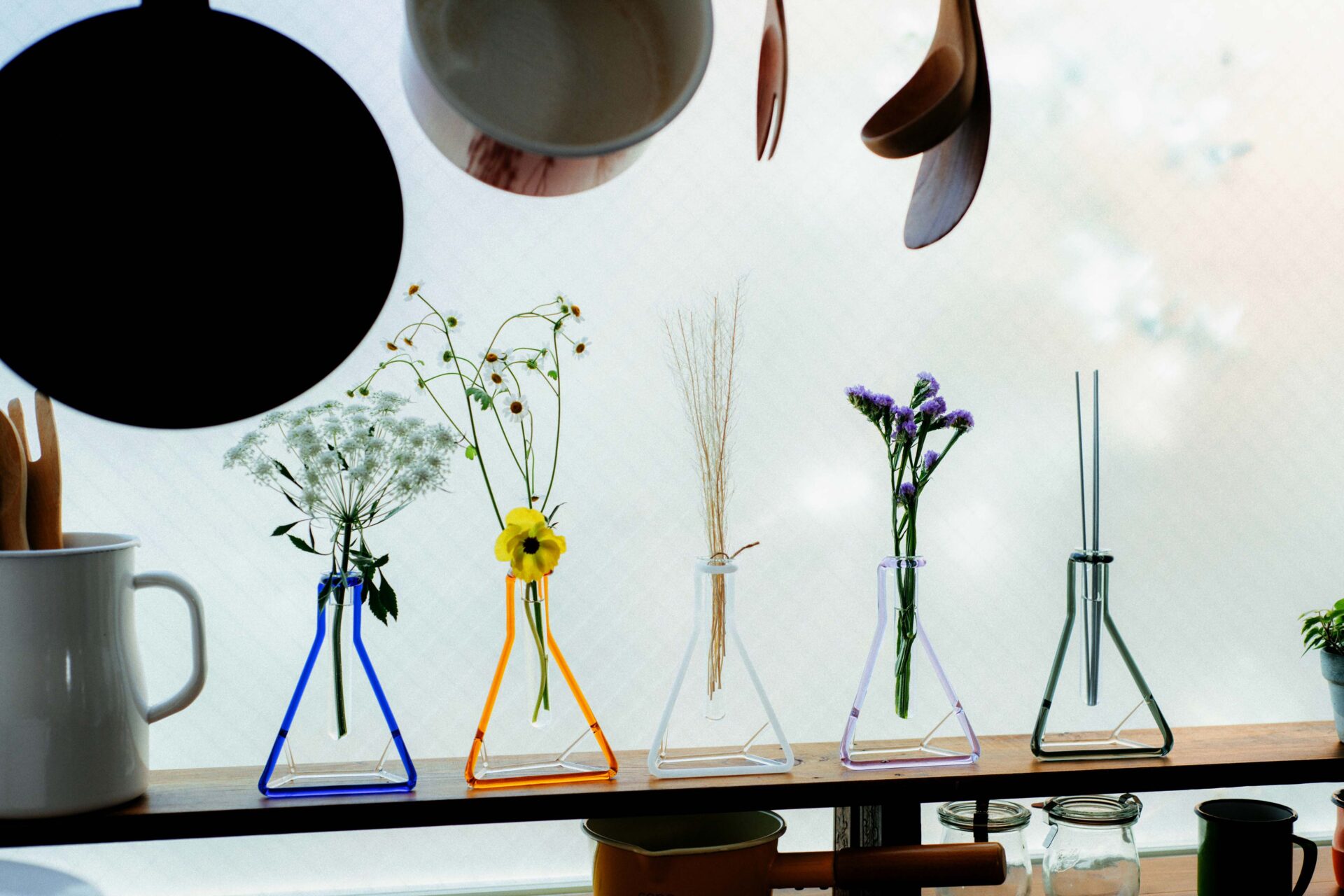 さんかくフラスコのかたちを色のついたガラスでかたどったデザインの花瓶「ガラスベースフレーム さんかく」が５つ、キッチンの窓辺に飾られている写真。それぞれに小さな花がいけられています