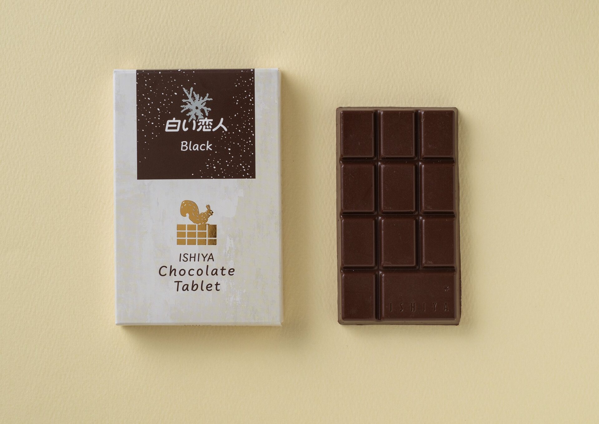 「チョコレートタブレット（白い恋人ブラック）」の白をベースにし、エゾリスのシルエットが描かれたパッケージの箱となかのチョコレートの写真。こちらはミルクチョコレートで、手でわれるように凹凸がついています
